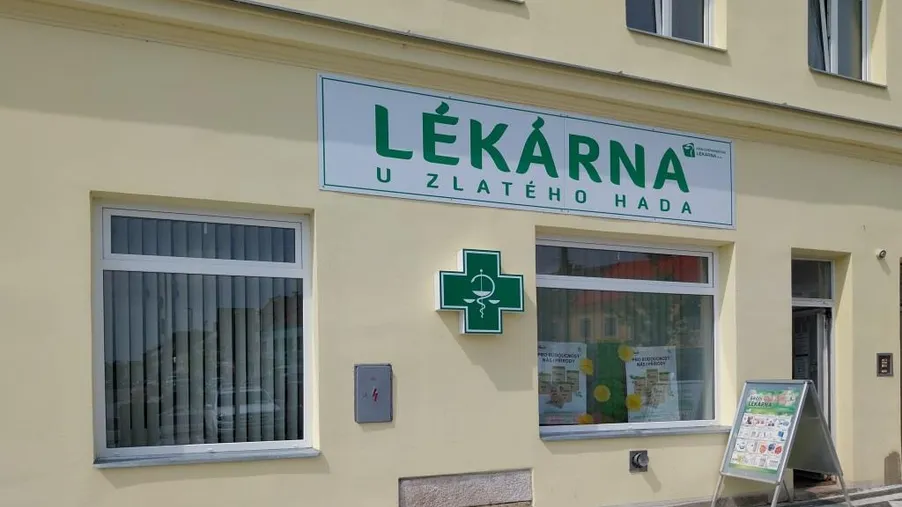 Pohled na fasádu lékárny U Zlatého hada s nápisem, vývěsním štítem a zeleným křížem, v historické budově s dlažbou před vchodem.